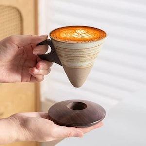 May include: A unique ceramic coffee cup with a brown handle and a conical shape, filled with latte art. The cup is held above a matching wooden coaster. The cup has a beige and brown striped design.