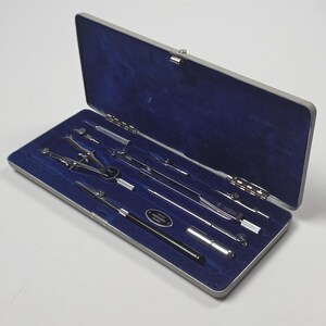 Può includere: Un set da disegno in metallo color argento in una custodia rettangolare con interno in velluto blu intenso. Il set include vari compassi in metallo, un tiralinee e un piccolo contenitore, con una targhetta metallica che riporta "rostfreier stahl".