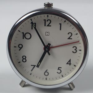 May include: A round analog alarm clock with a polished chrome metal frame and two small feet. The clock face is off-white with black Arabic numerals from 1 to 12, black hour and minute hands, a thin red second hand, and a small logo below the 12.