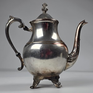 Op de afbeelding: Een gepolijste verzilverde theepot met een bolvormig lichaam, een gebogen handvat met zwarte accenten en een decoratieve tuit. Het deksel is voorzien van een knop en de basis rust op drie sierlijke, gebogen voetjes. Het oppervlak reflecteert licht.