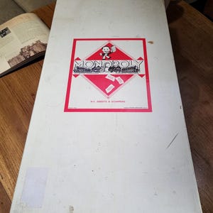 May include: A vintage Monopoly board game box. The box is white with a red and black logo featuring the Monopoly name and a cartoon character. The box has some wear and tear, suggesting it is an older edition of the classic board game.