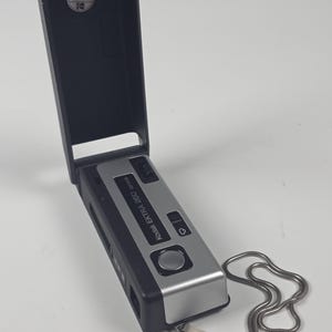 May include: A black and silver Kodak Ektra 260 camera with an open hinged cover. The camera features a silver faceplate with the text Kodak Ektra 260, a black shutter button, a small black switch, and a silver metal wrist strap attached to the side.