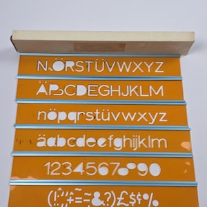 Könnte beinhalten: Ein Set aus sechs orangefarbenen Kunststoff-Schablonenstreifen mit blauen Metallkanten, untergebracht in einem rechteckigen Karton. Die Schablonen zeigen ausgeschnittene Buchstaben des Alphabets in Groß- und Kleinschreibung, Zahlen und Symbole.