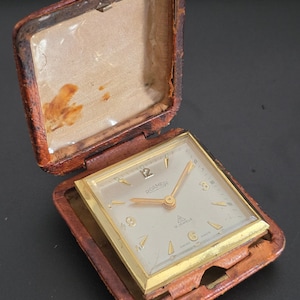 Puede incluir: Un reloj de viaje cuadrado Roamer con caja de metal dorado y esfera clara con manecillas e índices dorados. El reloj se encuentra en un estuche protector con bisagras cubierto de cuero marrón con un forro de tela de color beige.