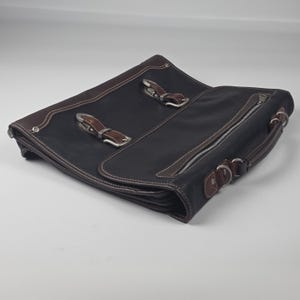 Puede incluir: Un maletín negro con ribetes de cuero marrón y herrajes de metal plateado. El bolso cuenta con dos correas frontales con hebillas, un asa superior y un bolsillo lateral con cremallera. El diseño incluye costuras visibles y anillas en forma de D.