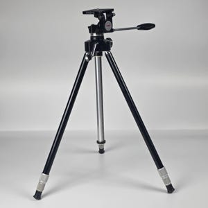 Może przedstawiać: Czarno-srebrny statyw fotograficzny Minette z trzema regulowanymi nogami zakończonymi gumowymi stopkami, centralną kolumną pionową oraz głowicą z uchwytem sterującym. Na górnej płytce montażowej widnieje nazwa marki Minette.