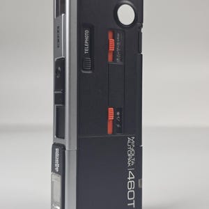 Puede incluir: Una cámara de bolsillo Minolta Autopak 460T plateada y negra con cuerpo rectangular, botón disparador blanco, interruptores deslizantes naranjas para teleobjetivo y ajustes de luz, y un flash integrado con la etiqueta Minolta Pocket.