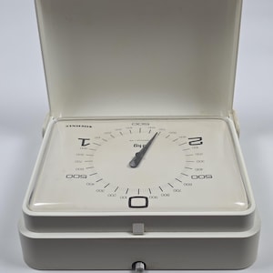 May include: A vintage off-white Soehnle kitchen scale with a hinged lid. The square face features a clear glass cover, a black needle, and a dial marked with numbers from 0 to 2 kg, with 500g increments clearly printed in black on the white surface.