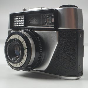 May include: A vintage silver and black OGA Germany camera featuring a Color Gotar 1:2.8/45 lens. The camera body has a textured black grip, a silver metal top plate, and a lens barrel with visible aperture and focus distance markings.