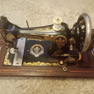 Può includere: Una macchina da cucire nera Gritzner Extra con intricati motivi floreali dorati, una ruota a manovella argentata e un rocchetto di filo bianco sulla parte superiore. La macchina è posizionata su una base rettangolare in legno con venature scure.