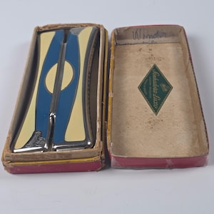 Könnte beinhalten: Eine Vintage-Mundharmonika mit blau-cremefarbener Emaille-Oberfläche liegt in einer abgenutzten Pappschachtel. Der Deckel der Schachtel zeigt ein rautenförmiges Etikett mit dem Text Sonhadora-Luxo Industria Brasileira und handschriftlicher Notiz.