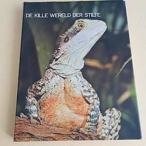 May include: A hardback book titled DE KILLE WERELD DER STILTE featuring a colour photograph of a lizard with textured grey and orange scales perched on a brown branch against a dark, blurred background. The lizard is looking towards the right.