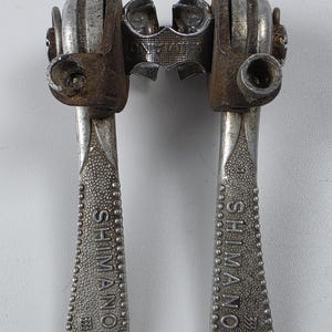 May include: A pair of vintage Shimano bicycle down tube shifters with a metallic silver finish and textured grip surfaces. The levers feature the Shimano brand name embossed on the front, with rusted mounting hardware and a central metal bracket.