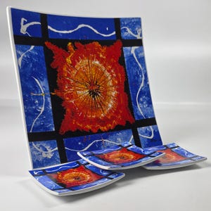May include: A large square plate and three smaller square plates featuring a vibrant design of a central red and orange sunburst pattern surrounded by a blue background with white wavy lines, all framed by thick black grid lines on a white base.
