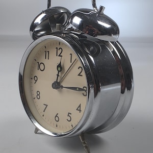 Könnte beinhalten: Ein verchromter Doppelglocken-Wecker mit cremefarbenem Zifferblatt, schwarzen arabischen Ziffern sowie schwarzen Stunden-, Minuten- und Sekundenzeigern. Die Uhr verfügt über einen Tragegriff, zwei Glocken und zwei kleine Metallfüße.