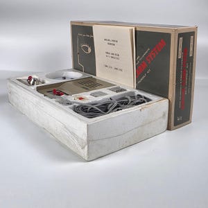Könnte beinhalten: Ein Do-it-yourself-Einbruchmeldeanlagen-Set in einem Karton mit Styroporeinsatz. Das Set enthält ein beiges Steuergerät, graue Elektrokabel, eine rote Lichtkomponente und eine Bedienungsanleitung mit dem Titel Burglar Alarm System.