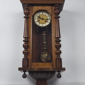 Könnte beinhalten: Eine braune Wanduhr aus Holz mit einem runden Zifferblatt, römischen Ziffern, schwarzen Zeigern und einem messingfarbenen Zentrum. Die Uhr wird von zwei vertikalen Säulen mit gedrechselten Details gerahmt und hat ein langes Pendel.