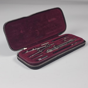 Könnte beinhalten: Ein schwarzes Etui mit Reißverschluss und einem tiefburgunderroten Samtfutter. Das Etui enthält ein Set aus silberfarbenen Metall-Zeicheninstrumenten, darunter Zirkel, Stechzirkel und kleine zylindrische Aufsätze in geformten Fächern.