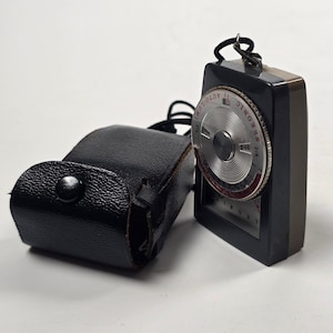 Può includere: Un esposimetro Sekonic Auto-Lumi nero con un quadrante circolare argentato che presenta scale numeriche e testo, abbinato a una custodia in pelle nera testurizzata con chiusura a bottone automatico e un sottile cinturino nero collegato.
