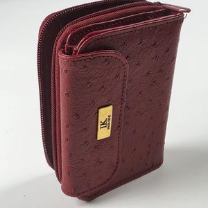 Könnte beinhalten: Ein dunkelrotes Bifold-Portemonnaie mit einer strukturierten Straußenleder-Optik. Das Portemonnaie verfügt über eine goldfarbene, rechteckige Metallplakette auf der Vorderklappe, die mit den Buchstaben IK und LAURA KENT graviert ist.