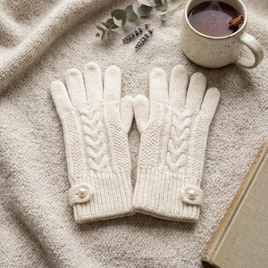 May include: A pair of cream-colored knit gloves featuring a vertical cable knit pattern on the back of the hand, a ribbed cuff, and a small tab detail at the wrist secured with a single white faux pearl button on each glove.