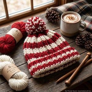 May include: A chunky knit beanie featuring horizontal stripes in deep red and cream, topped with a matching pom-pom. The hat is displayed alongside two skeins of yarn labeled Pine Cone Knits, wooden knitting needles, and a mug of hot cocoa.