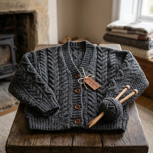 Puede incluir: Un cárdigan de punto trenzado en color gris carbón oscuro con cuello en V, mangas largas y cuatro botones marrones. Un ovillo de lana a juego con dos agujas de madera descansa sobre la prenda, junto a una etiqueta marrón Wildwool & Co.
