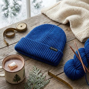 May include: A royal blue ribbed knit beanie with a folded cuff and a black rectangular tag featuring the white text ARCTIC WEAR. The beanie is placed on a wooden surface next to a measuring tape, a mug of hot cocoa, and blue yarn.
