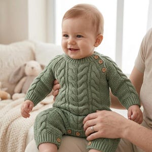 Può includere: Una tutina per neonati a maniche lunghe color verde salvia con un motivo a trecce verticali sul petto e sulle gambe, completata da maniche in maglia testurizzata. Il capo presenta uno scollo rotondo con tre piccoli bottoni in legno sulla spalla.
