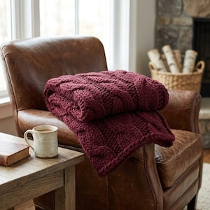 Può includere: Una coperta in maglia grossa color bordeaux intenso con un evidente motivo a trecce. Il filato spesso e strutturato crea un aspetto morbido e piegato mentre è drappeggiato su una poltrona in pelle marrone, offrendo un ricco contrasto cromatico.