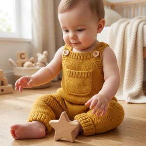 May include: A mustard yellow knitted romper featuring a front pocket, ribbed leg cuffs, and two light brown wooden buttons on the shoulder straps. The garment is made of a textured knit fabric and is worn by a baby sitting on a wooden floor.