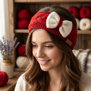 Puede incluir: Una diadema de punto roja con un patrón de trenzas y un lazo grande de color crema sujeto en el lateral. La diadema incluye una pequeña etiqueta rectangular marrón con la palabra Handmade impresa, situada cerca del lazo.