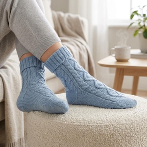 Puede incluir: Un par de calcetines de punto azul claro con un patrón de trenzas prominente que recorre verticalmente los lados. Los calcetines tienen un puño grueso y acanalado en la parte superior y están hechos de hilo texturizado, ofreciendo un aspecto suave.