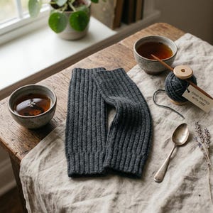 Puede incluir: Un par de calentadores de brazos de punto acanalado gris oscuro junto a dos cuencos de cerámica llenos de té, un carrete de madera con hilo gris oscuro, una cuchara de plata y una pequeña etiqueta con el texto Hand-knitted with care.