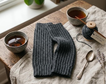 May include: A pair of dark gray ribbed knit arm warmers positioned next to two ceramic bowls filled with tea, a wooden spool with dark gray yarn, a silver spoon, and a small tag with the text Hand-knitted with care, all on a linen cloth.