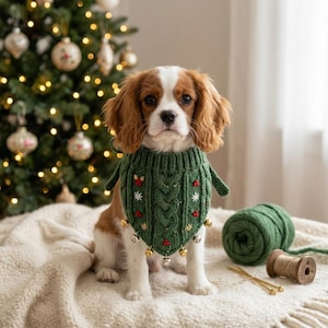 Op de afbeelding: Een Cavalier King Charles-spaniël draagt een donkergroene gebreide bandana voor huisdieren met een kabelpatroon, geborduurde hulstbessen en kleine witte sneeuwvlokken. De driehoekige bandana is versierd met diverse kleine gouden belletjes.