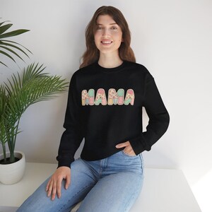 May include: Black sweatshirt featuring the word "MAMA" in a patchwork design. The letters are decorated with pink, blue, and yellow patterns. The long-sleeved top is worn with blue jeans.