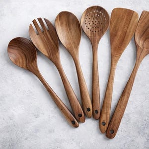 Puede incluir: Un conjunto de seis utensilios de cocina de madera, que incluyen una cuchara, una cuchara ranurada, una esp&aacute;tula y un tenedor para servir. Los utensilios son de madera de color marr&oacute;n claro y est&aacute;n dispuestos sobre una superficie gris claro.