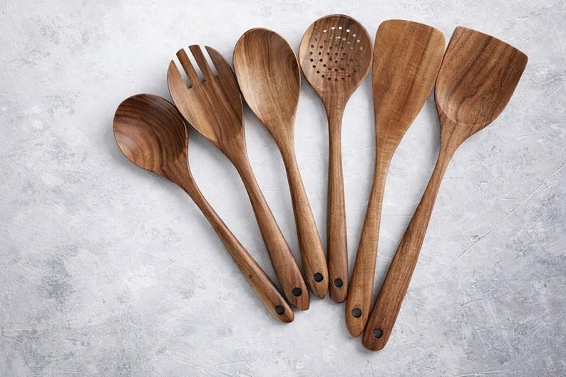 Puede incluir: Un conjunto de seis utensilios de cocina de madera, que incluyen una cuchara, una cuchara ranurada, una esp&aacute;tula y un tenedor para servir. Los utensilios son de madera de color marr&oacute;n claro y est&aacute;n dispuestos sobre una superficie gris claro.