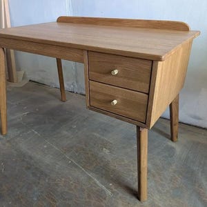 Peut inclure: Un bureau en bois brun clair doté d'un plateau rectangulaire avec un rebord arrière surélevé, quatre pieds fuselés et deux tiroirs superposés sur le côté droit. Chaque tiroir est muni d'un petit bouton de poignée cylindrique en bois clair.