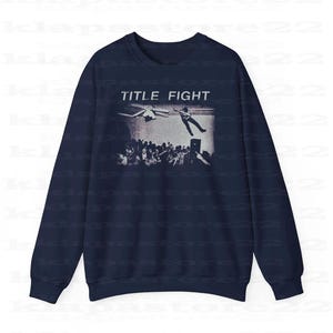 Peut inclure: Sweat-shirt bleu marine avec un motif noir et blanc repr&eacute;sentant une foule, une personne qui tombe et un avion. Le texte "TITLE FIGHT" est imprim&eacute; au-dessus du motif en blanc.