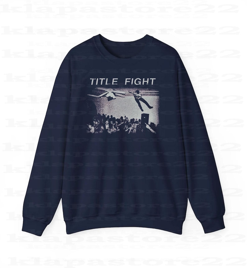 Peut inclure: Sweat-shirt bleu marine avec un motif noir et blanc repr&eacute;sentant une foule, une personne qui tombe et un avion. Le texte "TITLE FIGHT" est imprim&eacute; au-dessus du motif en blanc.
