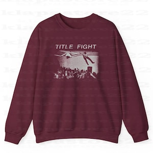 Peut inclure: Sweat-shirt bordeaux avec une impression graphique blanche. Le graphisme pr&eacute;sente les mots "TITLE FIGHT" au-dessus d'une foule de personnes regardant une personne tomber d'une sc&egrave;ne. Le sweat-shirt a des manches longues et un col rond.