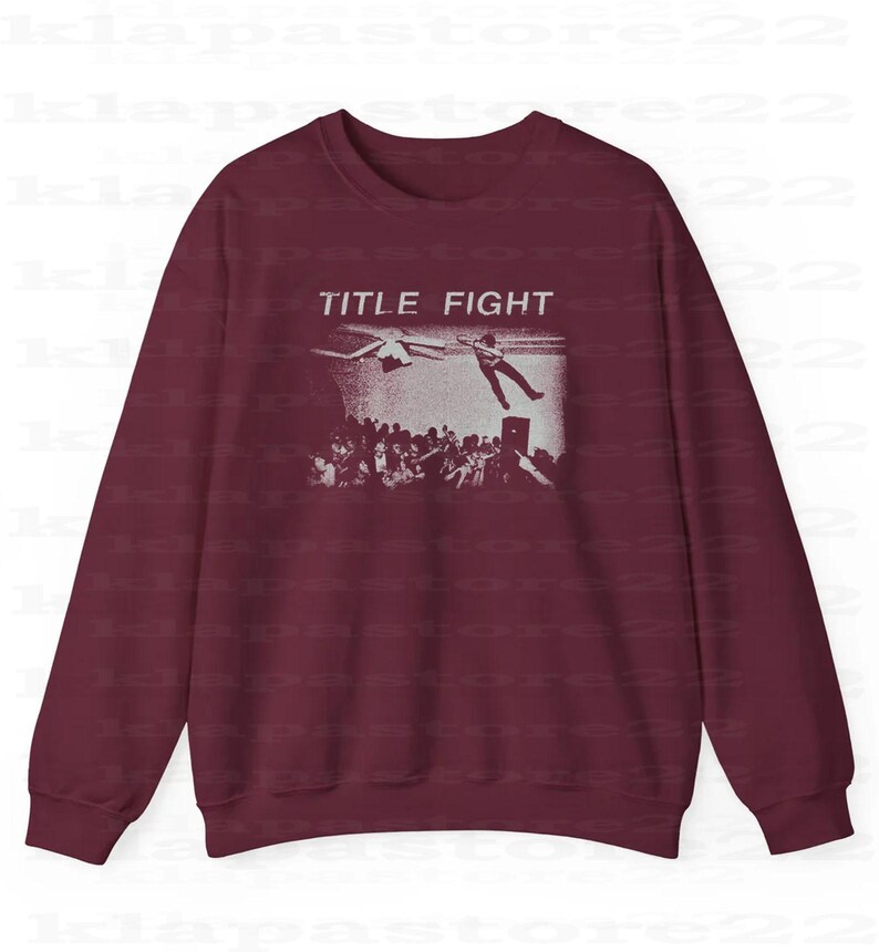 Peut inclure: Sweat-shirt bordeaux avec une impression graphique blanche. Le graphisme pr&eacute;sente les mots "TITLE FIGHT" au-dessus d'une foule de personnes regardant une personne tomber d'une sc&egrave;ne. Le sweat-shirt a des manches longues et un col rond.