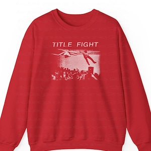 Peut inclure: Un sweat-shirt rouge avec un motif blanc. Le motif affiche le texte "TITLE FIGHT", une personne tombant d'une sc&egrave;ne, une foule et un h&eacute;licopt&egrave;re. V&ecirc;tement d&eacute;contract&eacute;.