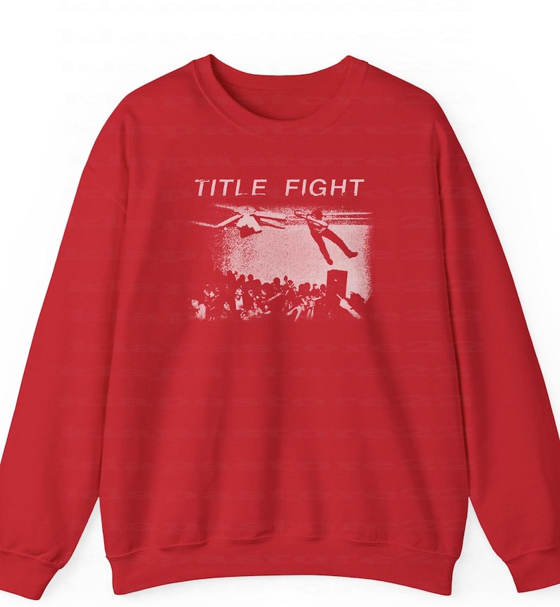 Peut inclure: Un sweat-shirt rouge avec un motif blanc. Le motif affiche le texte "TITLE FIGHT", une personne tombant d'une sc&egrave;ne, une foule et un h&eacute;licopt&egrave;re. V&ecirc;tement d&eacute;contract&eacute;.