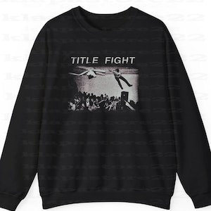 Peut inclure: Sweat-shirt noir avec un graphisme en niveaux de gris repr&eacute;sentant une sc&egrave;ne de concert. Les mots "TITLE FIGHT" sont au-dessus de l'image. Le graphisme montre une personne sautant d'une sc&egrave;ne avec une foule en dessous.