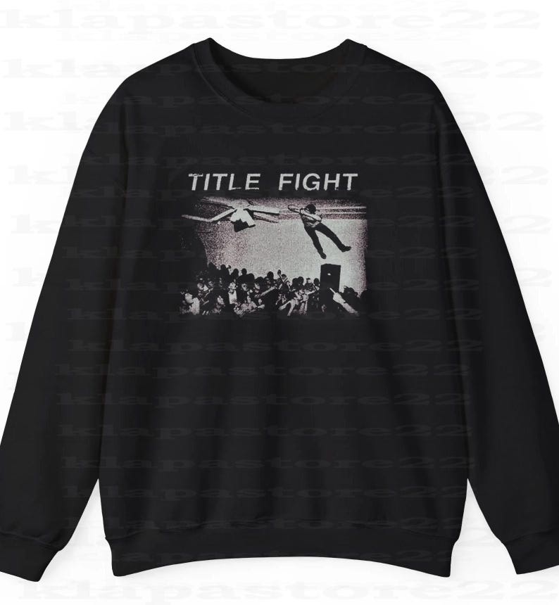 Peut inclure: Sweat-shirt noir avec un graphisme en niveaux de gris repr&eacute;sentant une sc&egrave;ne de concert. Les mots "TITLE FIGHT" sont au-dessus de l'image. Le graphisme montre une personne sautant d'une sc&egrave;ne avec une foule en dessous.