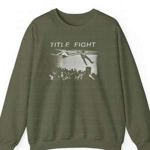 Peut inclure: Un sweat-shirt vert olive avec une impression graphique blanche. Le graphisme pr&eacute;sente les mots "TITLE FIGHT" au-dessus d'une foule de personnes et d'une personne tombant du ciel. Le sweat-shirt a des manches longues et un col rond.