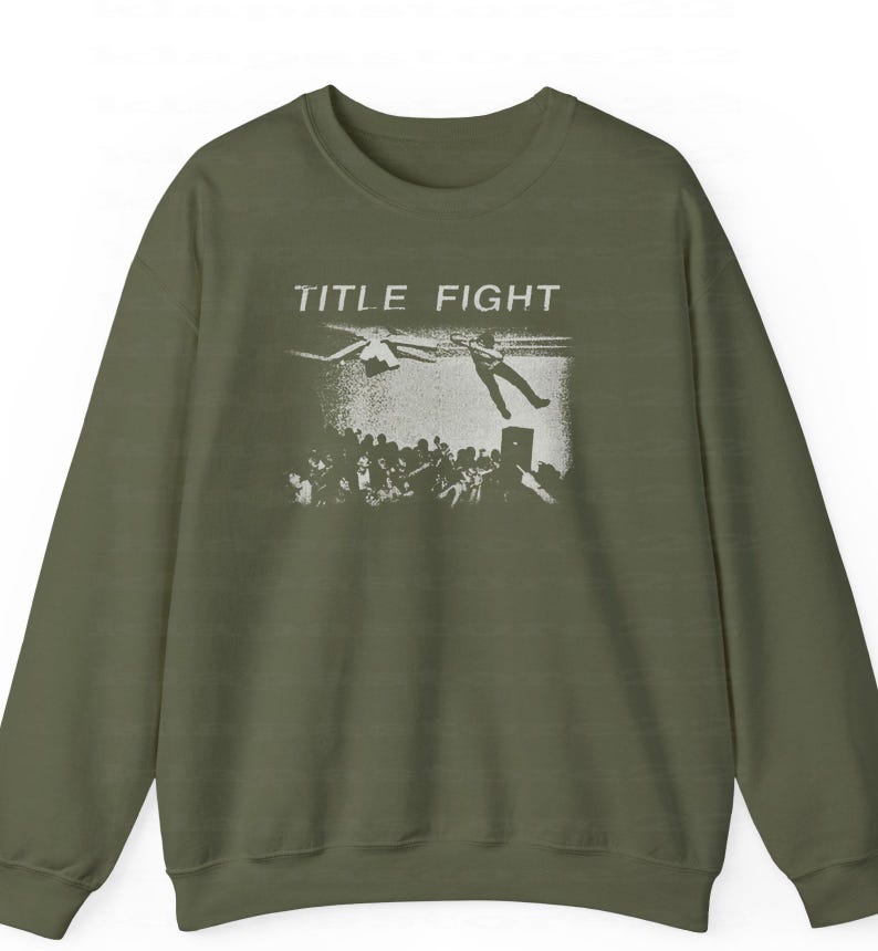 Peut inclure: Un sweat-shirt vert olive avec une impression graphique blanche. Le graphisme pr&eacute;sente les mots "TITLE FIGHT" au-dessus d'une foule de personnes et d'une personne tombant du ciel. Le sweat-shirt a des manches longues et un col rond.